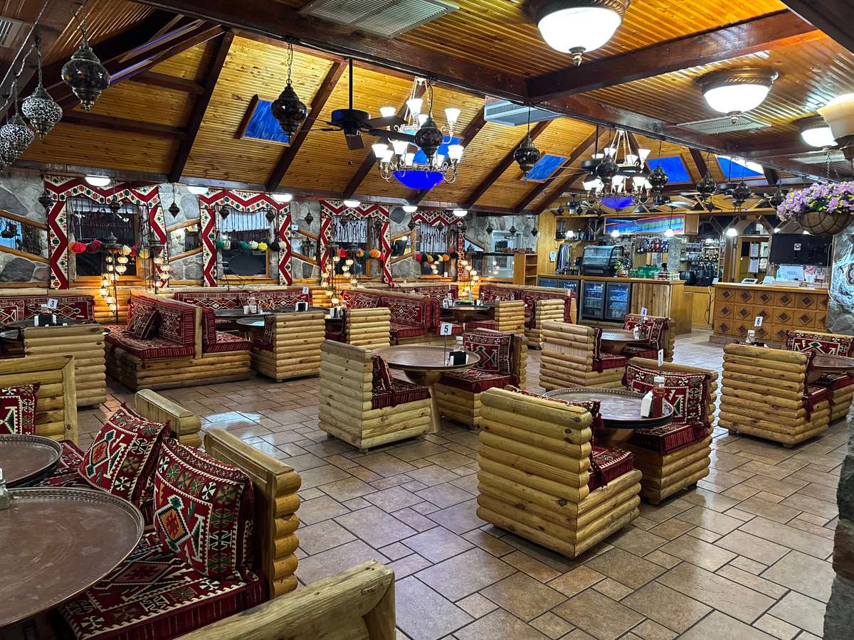 Cozy Mediterranean restaurant with wooden log seating, red patterned cushions, hanging lanterns, and a rustic stone-accented interior.