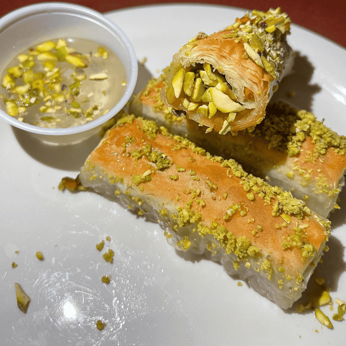 Flaky pistachio-filled baklava rolls drizzled with syrup, served with a side of crushed pistachio syrup for dipping.