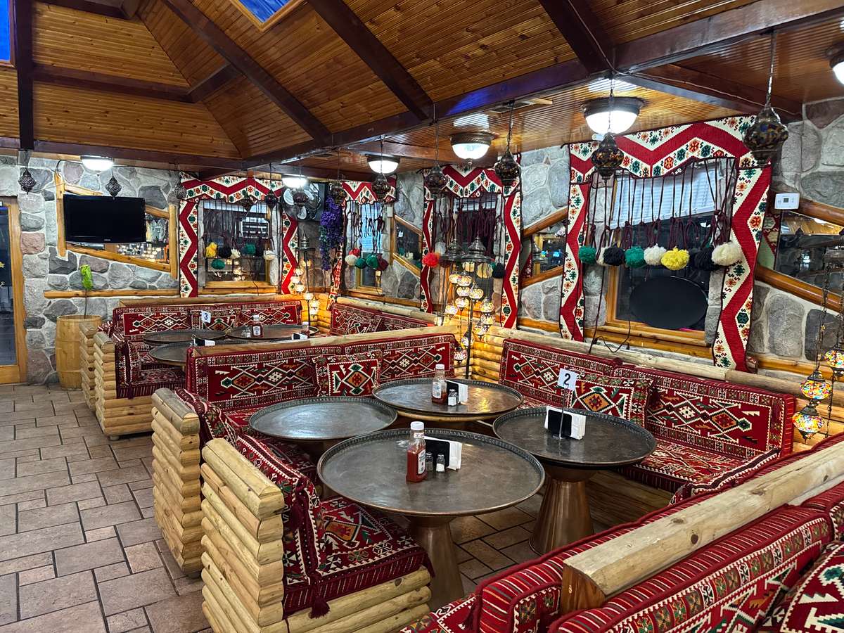 Warm Mediterranean-style dining space with wooden log seating, red patterned cushions, hanging lanterns, and stone walls for a cozy ambiance.