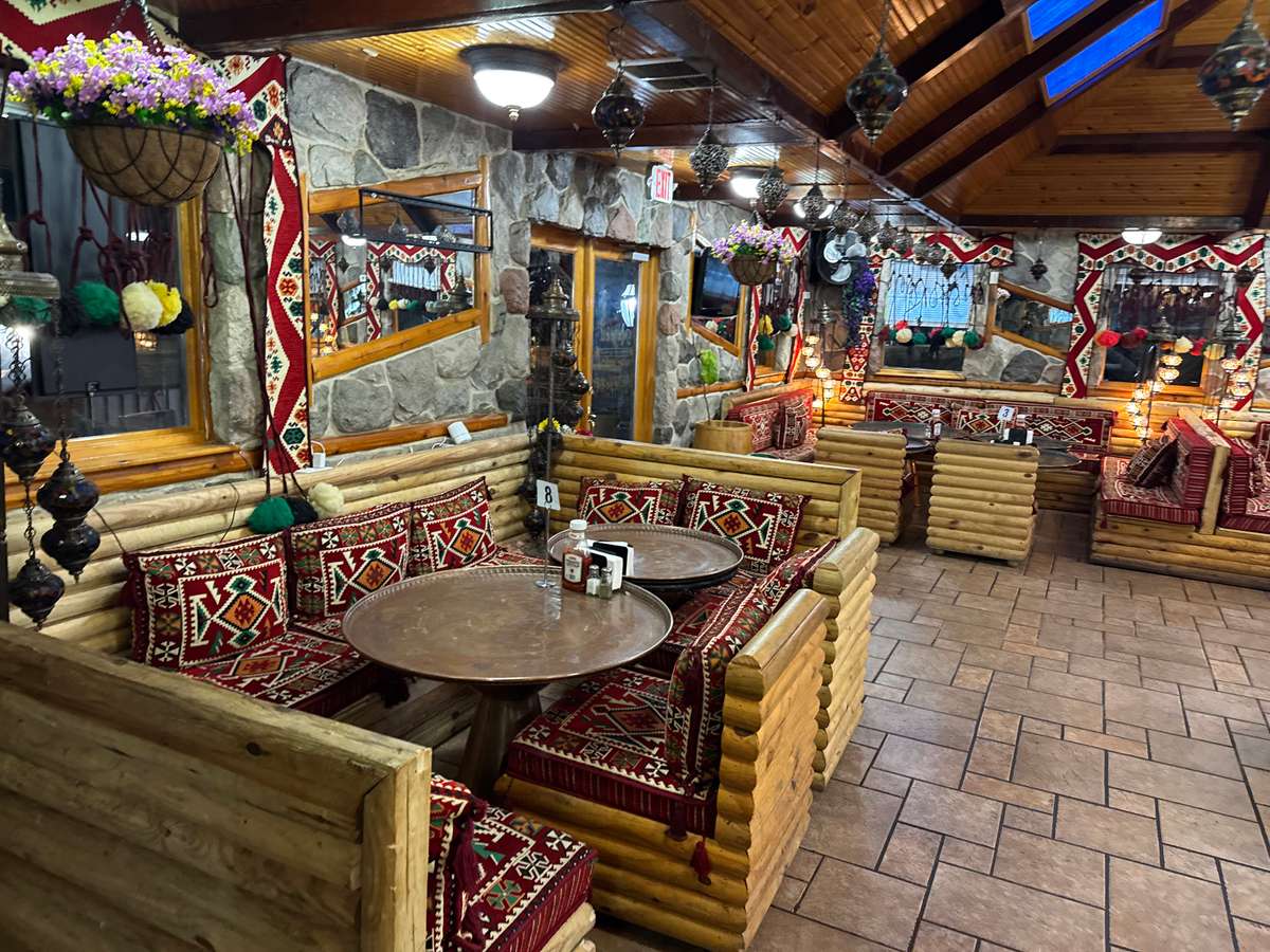 Cozy Mediterranean restaurant interior with wooden log seating, patterned red cushions, hanging lanterns, and stone walls for a warm atmosphere.