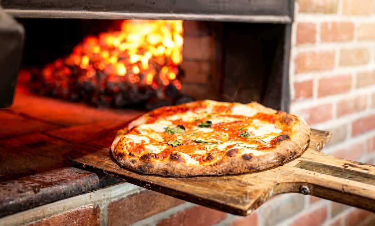 Fresh pizza on a wooden peel being placed into a brick wood-fired oven.