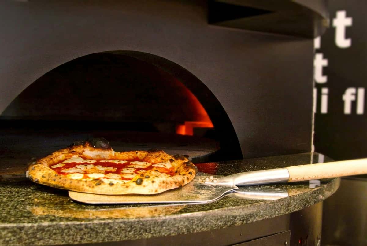 Pizza baking inside a commercial pizza oven with glowing heat in the background.