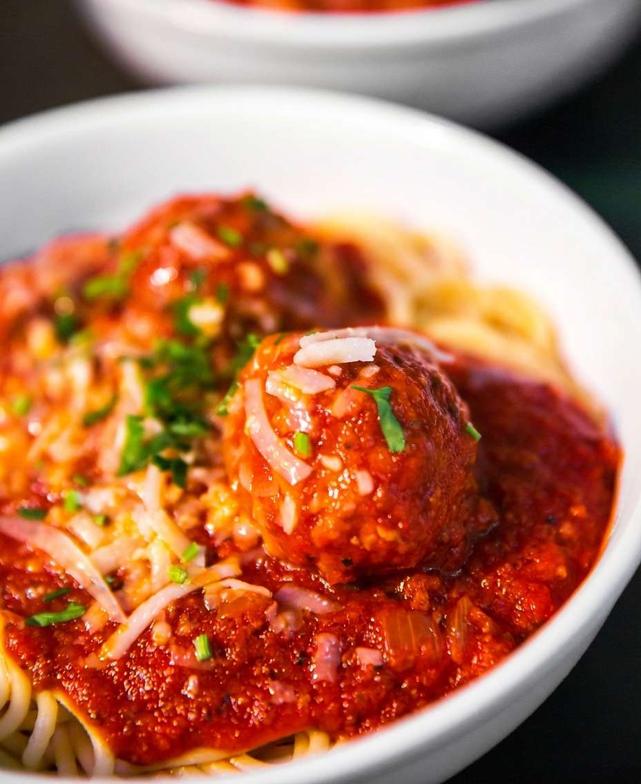 Bowl of spaghetti with marinara sauce and a large meatball topped with grated cheese and herbs.