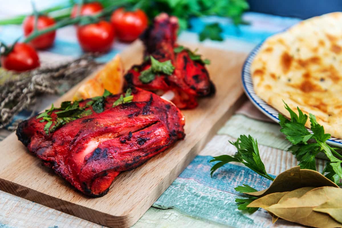 Grilled tandoori chicken served on a wooden board with herbs and flatbread.