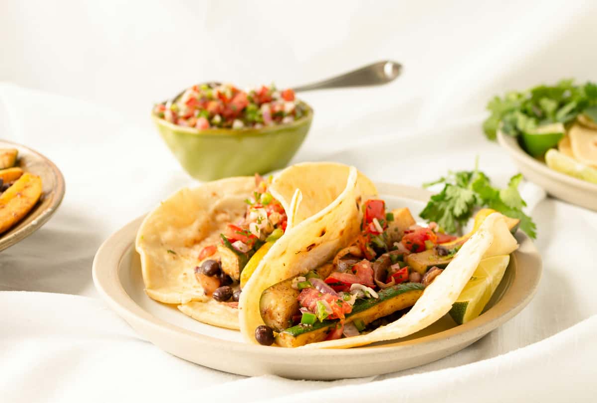 Soft flour tortillas filled with sautéed zucchini, black beans, and fresh pico de gallo, served with lime wedges and a side of salsa—perfect for a vibrant vegetarian taco dish.