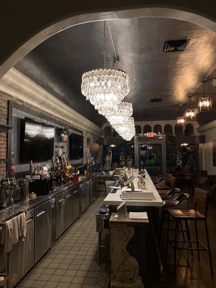 Elegant bar interior with crystal chandeliers, high stools, and stainless steel counters creating a warm evening ambiance.