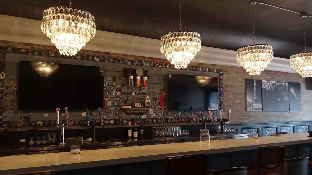 Bar with crystal chandeliers, two large TVs, and walls decorated with colorful patches above beer taps and wine bottles.