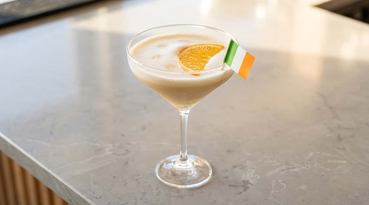 A white foamy cocktail with an orange slice and an Irish flag pick, adorned with small heart designs.