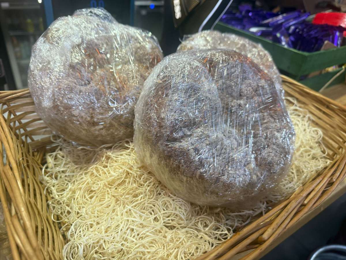 Two large round food items (possibly cakes, puddings, or dough-based desserts) wrapped tightly in plastic wrap, placed in a wicker basket on top of thin noodle-like decorative strands.