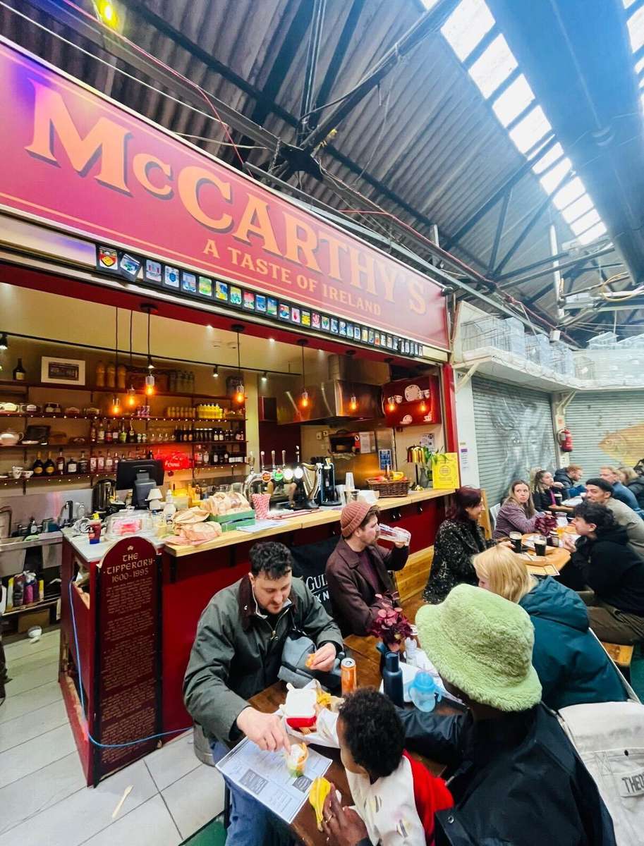A busy food stall called "McCarthy's – A Taste of Ireland" inside an indoor market, with a bar counter serving drinks and food while groups of people sit at long tables eating, drinking, and socializing.