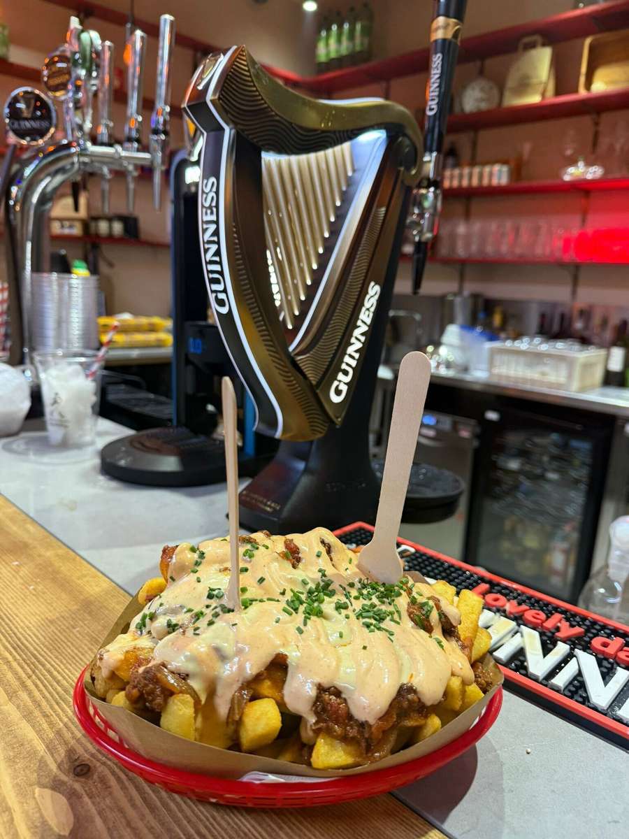Loaded fries topped with meat and melted creamy sauce, garnished with chopped chives, served on a plate at a bar counter with Guinness beer taps in the background.