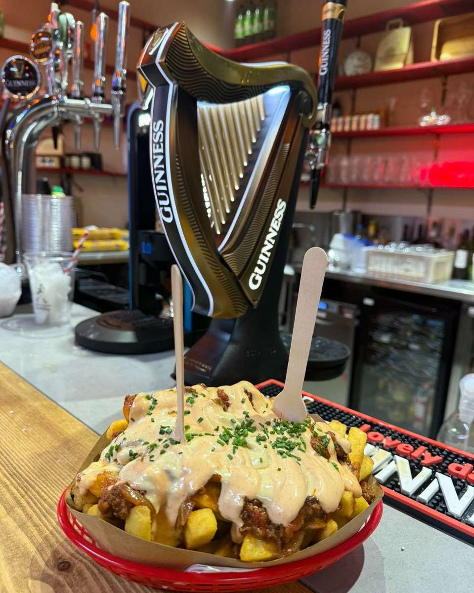Loaded cheesy fries topped with savory meat and creamy sauce, garnished with chopped chives, served at the bar in front of a Guinness tap