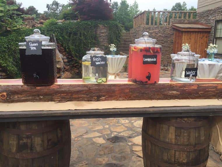 An outdoors photo of a decorated wooden board on barrels having beverages in glass pots.