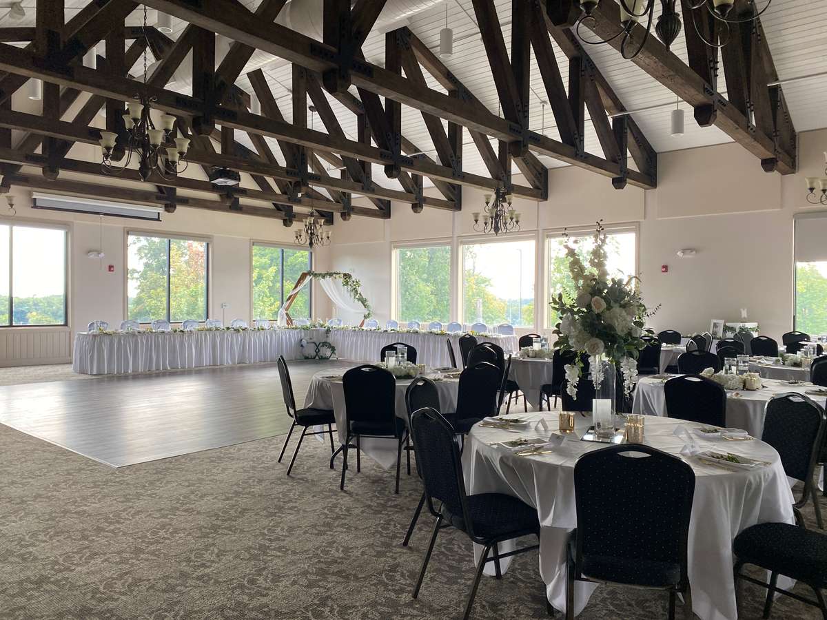 A room with carpeting and a small section of laminate flooring. It's set up with a head table. There are round tables set up to the right. All of the windows are open.