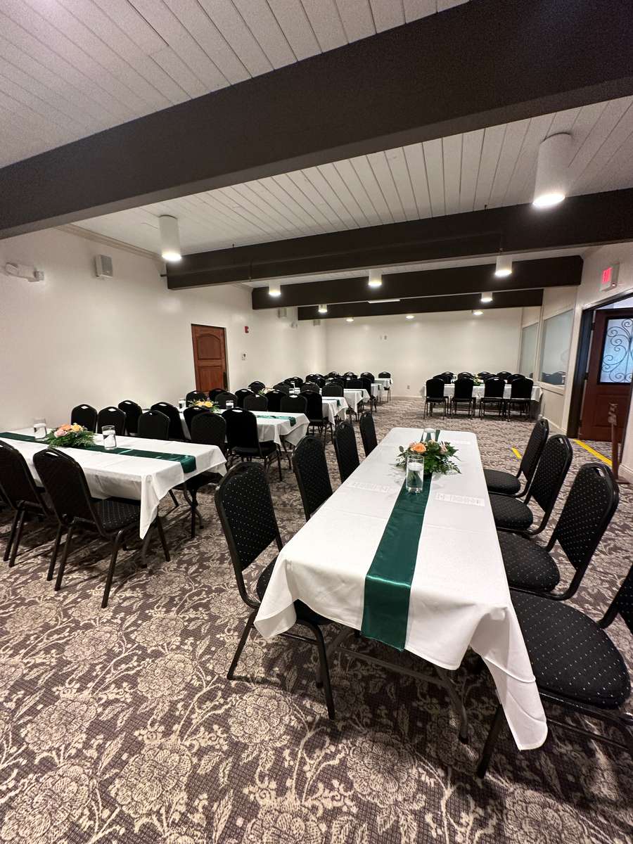 A smaller room set up with rectangular tables. The tables have white tablecloths, a green runner, and a small floral centerpiece.