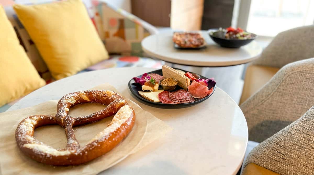 Soft pretzel with coarse salt on paper, charcuterie board with meats and cheese, and a small pizza on tables in a lounge setting.