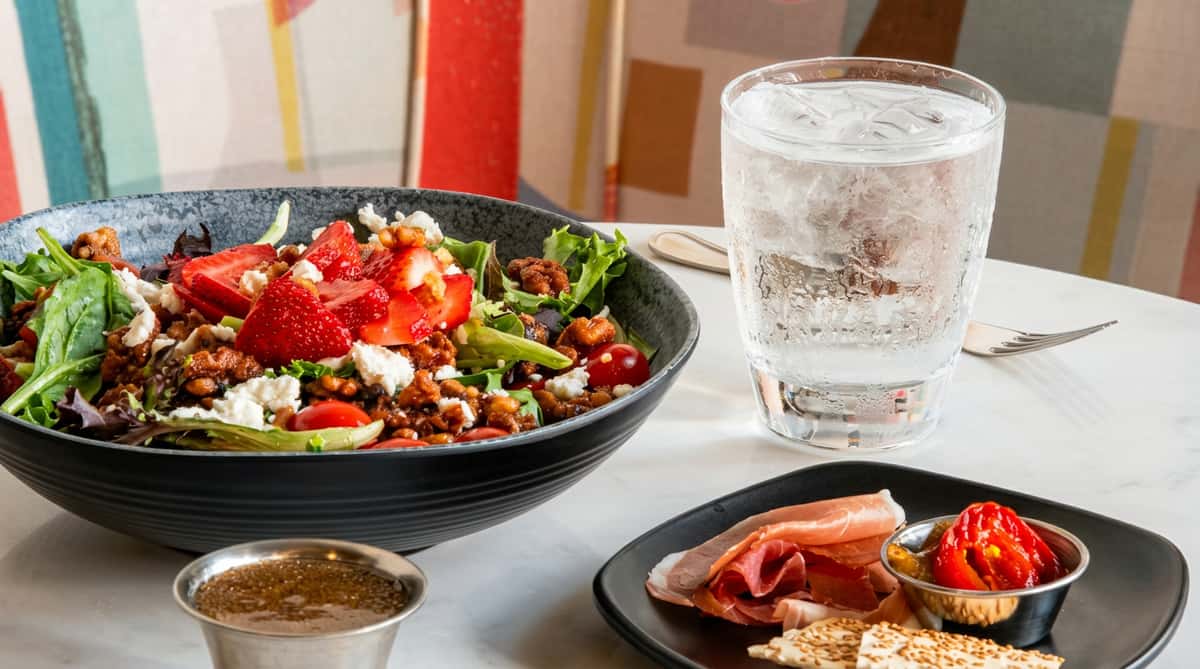 Salad with strawberries, feta, walnuts, leafy greens, tomatoes, prosciutto, and roasted red peppers. A glass of iced water.