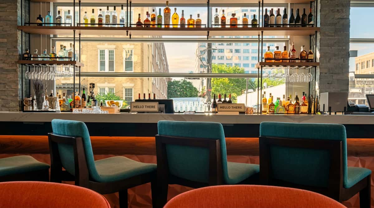Stylish bar interior with teal bar stools and orange chairs in the foreground, looking out to city buildings.