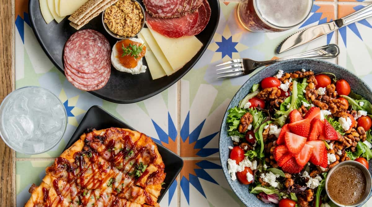 Charcuterie board with meats, cheeses, mustard, and crackers, BBQ flatbread, and a salad with strawberries, nuts, and feta.
