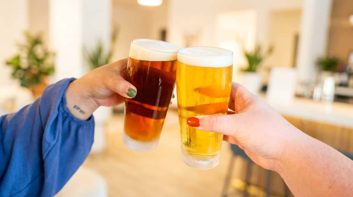 Two hands clinking tall glasses of beer, one amber and one golden, with foamy heads.