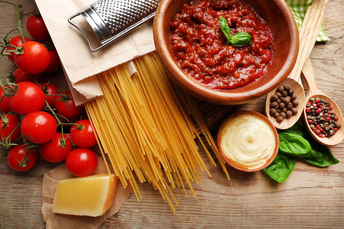 baby tomatoes, block of cheese, uncooked noodles, sauce in a bowl and a cheese grater