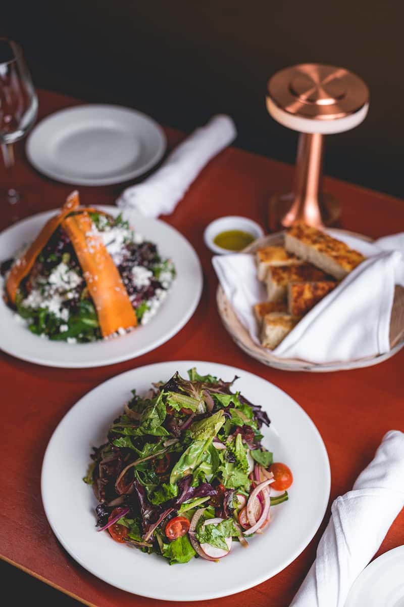 Overhead view of fresh Italian salads, roasted vegetables, bread, and appetizers arranged on a dining table.