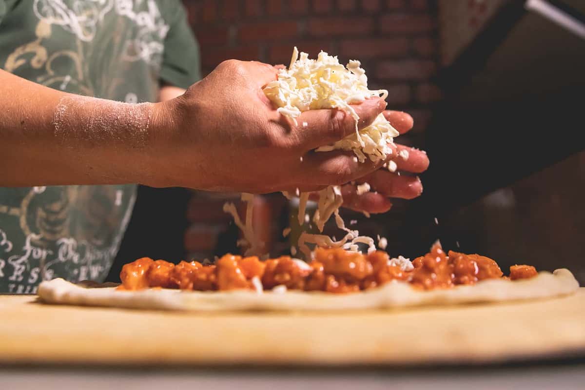 Chef grating fresh Parmesan cheese over a pizza topped with tomato sauce and Italian ingredients, highlighting freshly prepared cuisine at Pasta Lovers Trattoria in Midtown Manhattan, NYC.