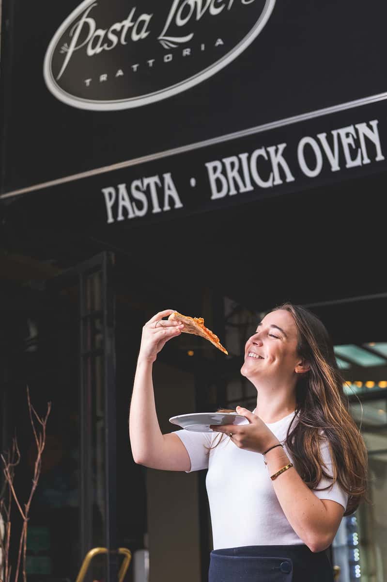 Woman enjoying a slice of Italian pizza outside Pasta Lovers Trattoria in Midtown Manhattan, NYC, beneath the restaurant's Pasta and Brick Oven sign.