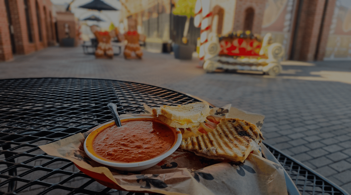 Tomato soup in a white bowl with a grilled cheese sandwich cut in half on a black metal table, against a blurred street background.
