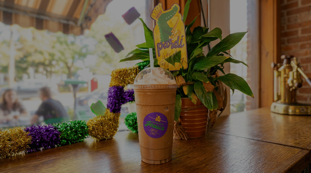 Frosted coffee drink with whipped cream and Louisiana Jeaux Coffee Cafe sticker on a wooden table with Mardi Gras decorations.