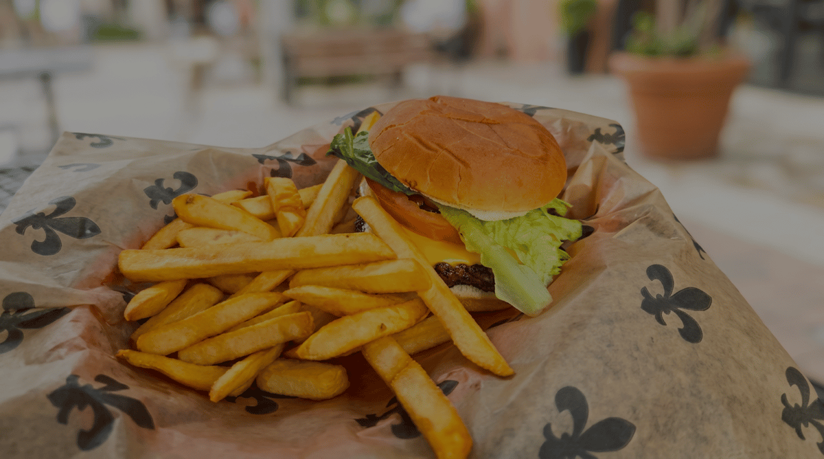 Cheeseburger with fresh lettuce, tomato, pickles, and melted cheese served with crispy French fries on a paper-lined basket in a casual outdoor dining setting.