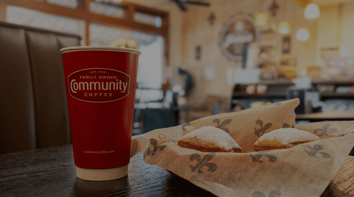 A red "Community Coffee" cup sits next to a basket of two beignets dusted with powdered sugar, in a cafe setting.