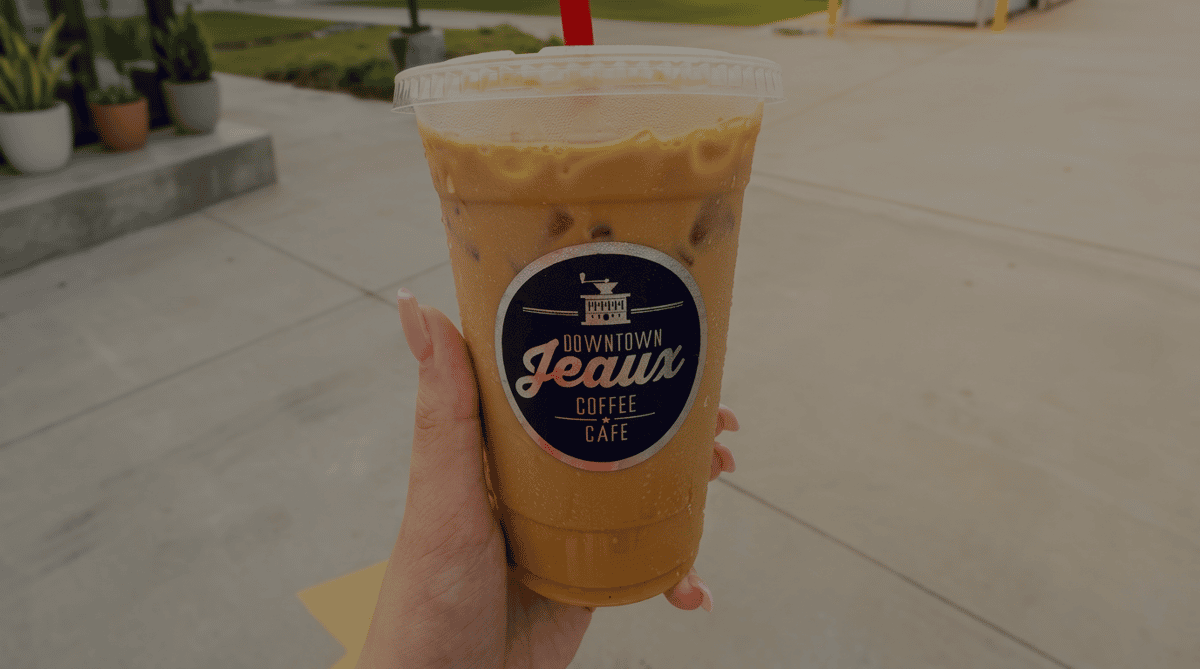 Hand holding an iced coffee from Downtown Jeaux Coffee Café in a clear cup with a red straw, enjoyed outdoors on a sunny day.