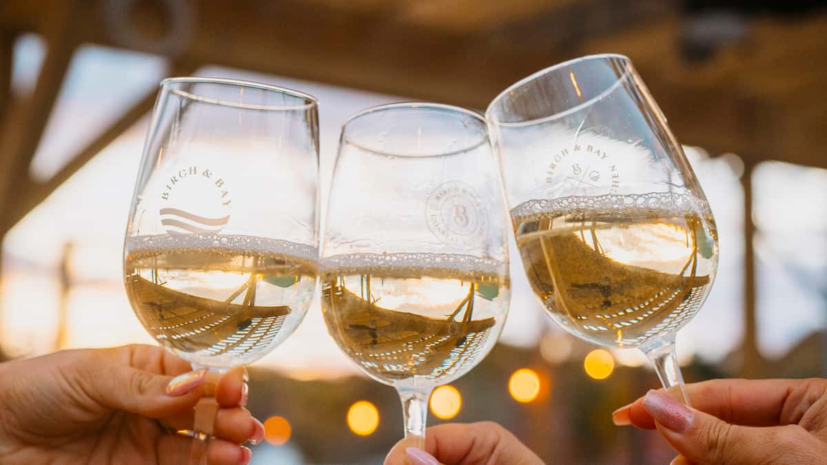 Three hands toasting with glasses of chilled white wine at sunset on an outdoor patio, with golden hour reflections and bokeh lights creating a warm, coastal ambiance — perfect for beachside restaurants, wine bars, or romantic dining settings.