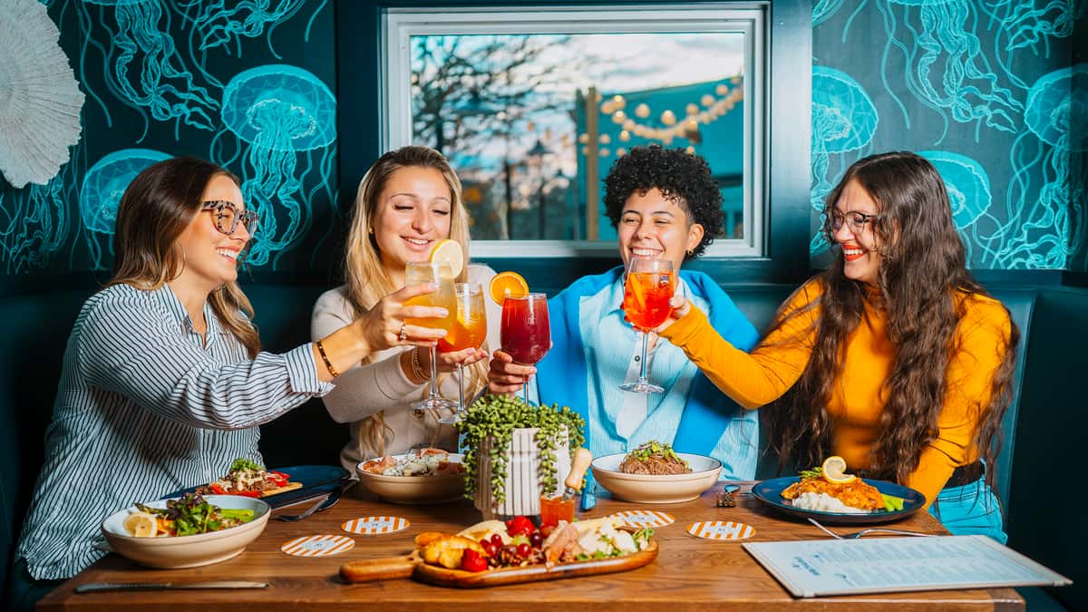 Group of friends toasting with colorful cocktails over a vibrant spread of salads, grain bowls, and shared appetizers at a trendy, modern restaurant with ocean-inspired decor — a lively dining scene perfect for social gatherings and weekend brunch vibes.