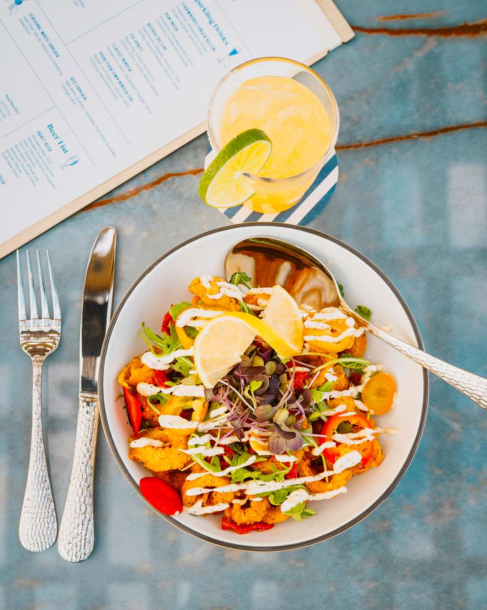 Crispy calamari bowl garnished with microgreens, lemon wedges, and drizzled aioli, served with roasted peppers and fresh herbs, accompanied by a citrus cocktail with lime — a vibrant coastal-inspired dish perfect for seafood restaurants and beachside dining menus.