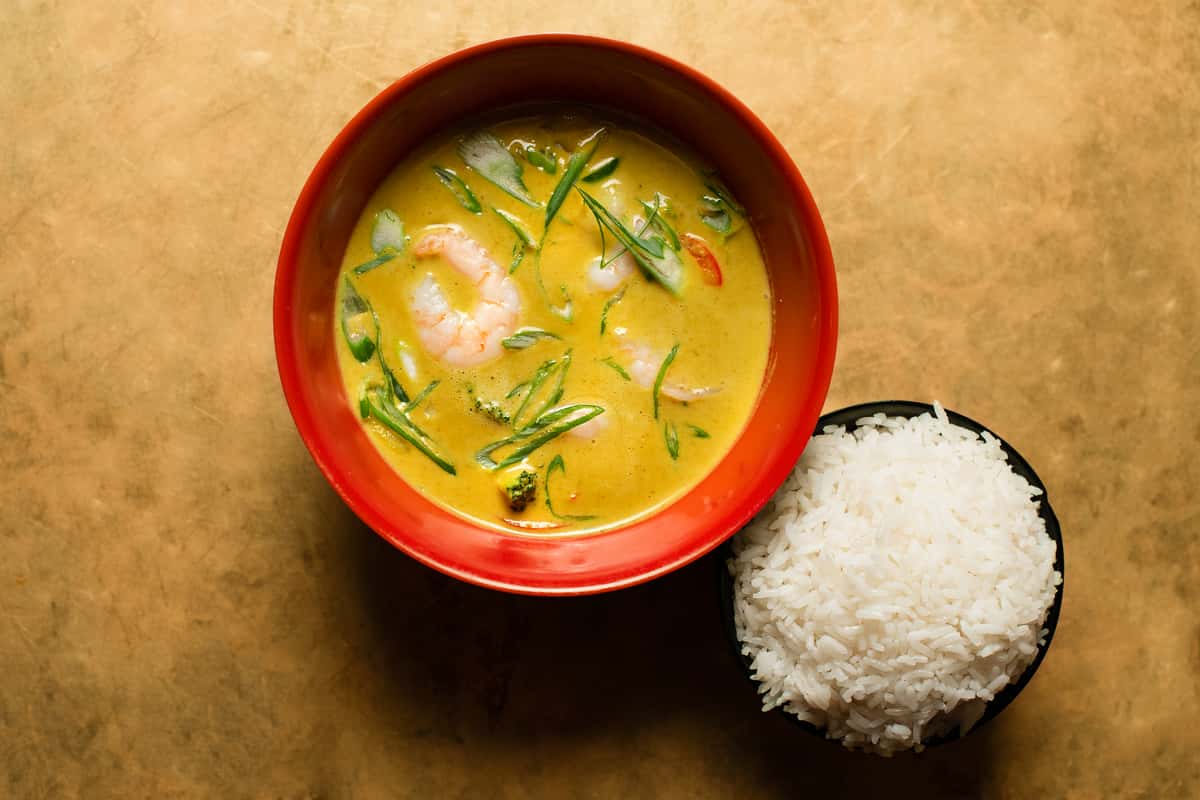 Creamy Thai-style shrimp coconut curry with fresh herbs and vegetables, served in a vibrant red bowl alongside steamed white rice in a modern Asian restaurant setting