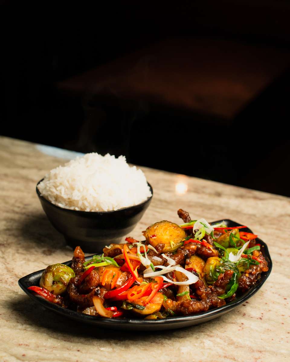 Spicy Asian stir-fry with crispy beef, roasted Brussels sprouts, and sliced red chilies, garnished with fresh scallions and served with a side of steamed white rice in a modern restaurant setting