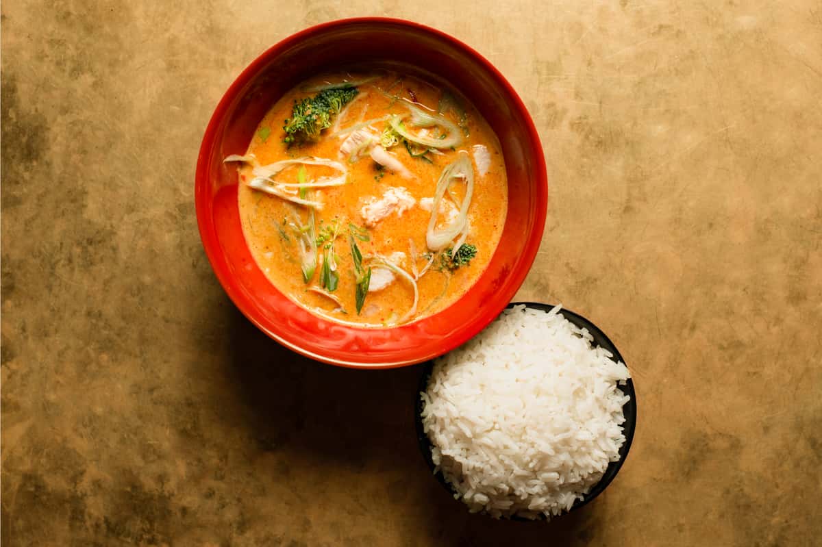 Creamy coconut curry soup with fresh vegetables and herbs, served in a vibrant red bowl alongside a side of steamed white rice in a modern Asian restaurant setting
