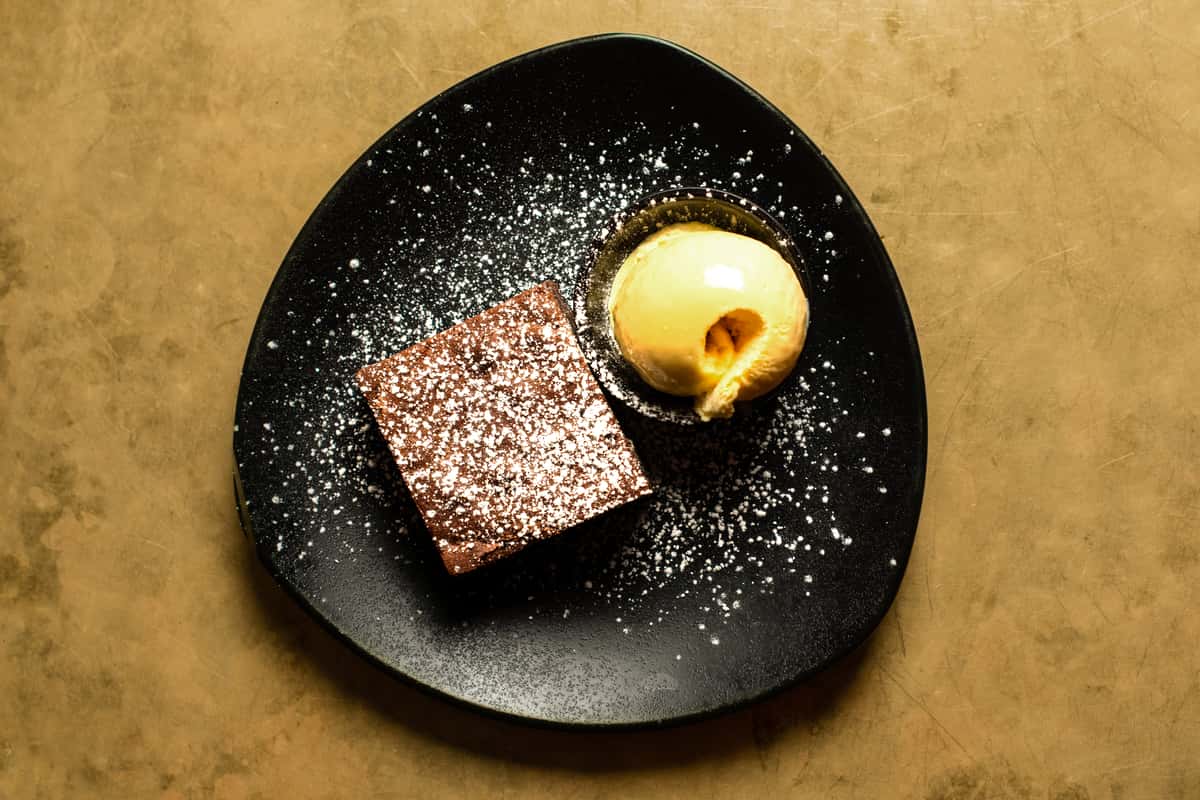 Decadent chocolate brownie dusted with powdered sugar, served with a scoop of vanilla ice cream on a modern black plate in a stylish restaurant setting
