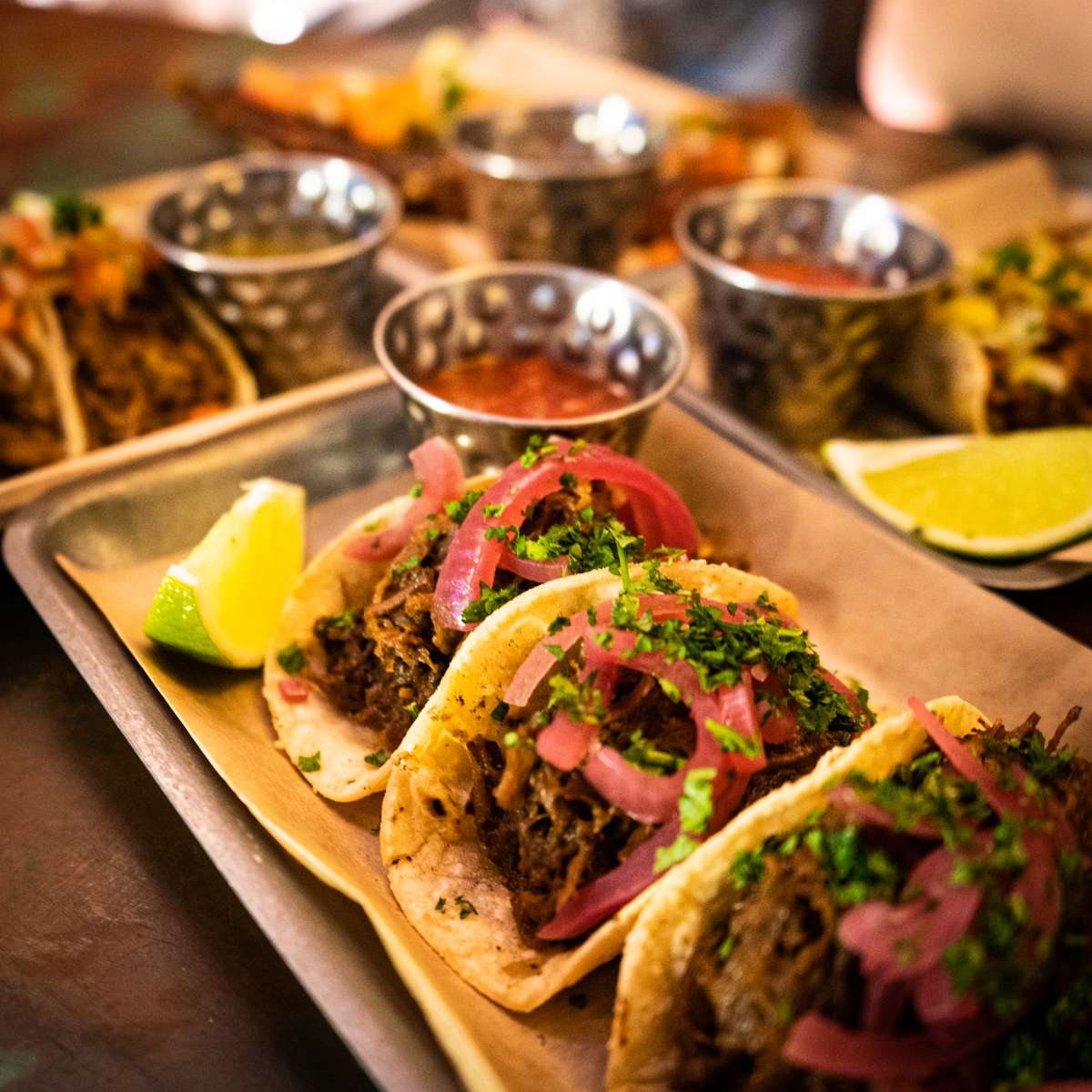 Three street tacos filled with seasoned meat, topped with pink onions and fresh cilantro, served on a tray with lime wedges and small bowls of salsa in the background.
