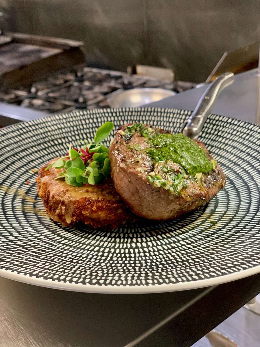 Juicy steak with green sauce and a breaded side on a patterned plate served at KP's Kitchen.