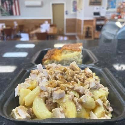 Macaroni and cheese topped with diced chicken and savory gravy, served in a black takeout container with a slice of golden cornbread in the background.