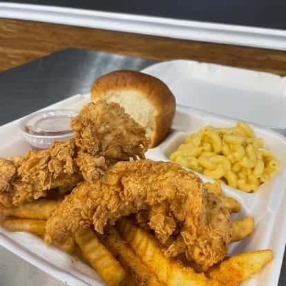 Takeout plate with crispy fried chicken tenders, seasoned fries, macaroni and cheese, a soft roll, and a side of dipping sauce.