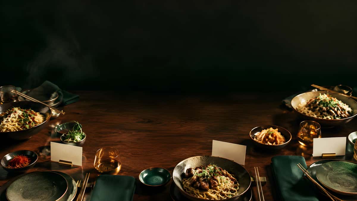 Asian noodle dishes with glazed meat, fresh greens, chili garnish, and elegant table settings in a moody restaurant