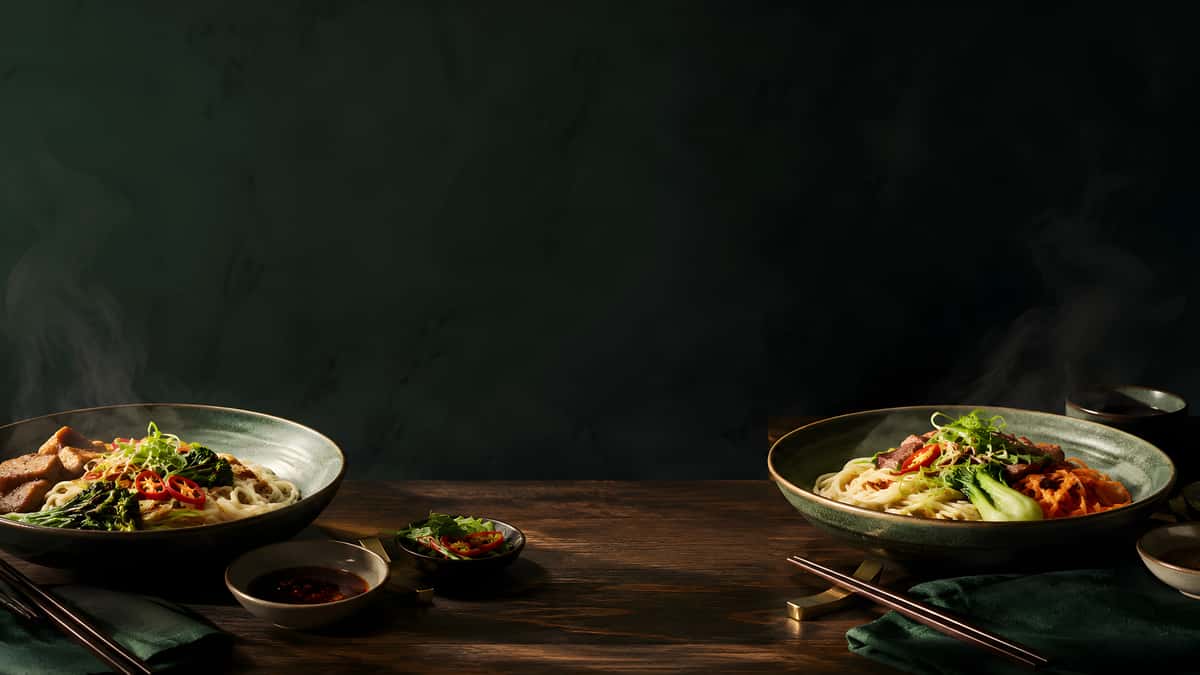 Steaming Asian noodle bowls with sliced meat, bok choy, fresh vegetables, chili peppers, and dipping sauces