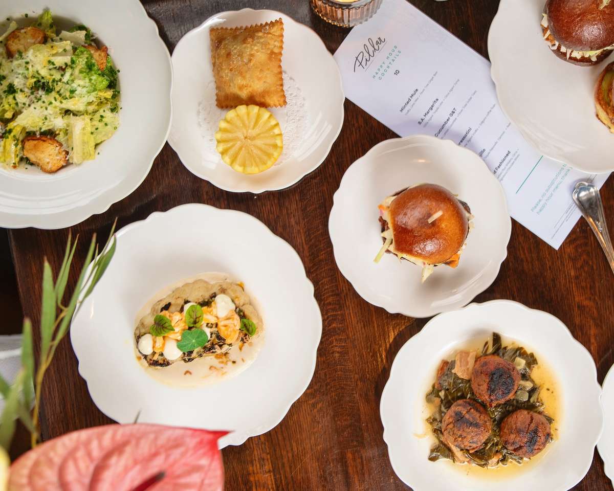 Elegant flat lay of small plates featuring sliders, empanadas, meatballs with greens, Caesar salad, and creamy vegetable-topped dish.