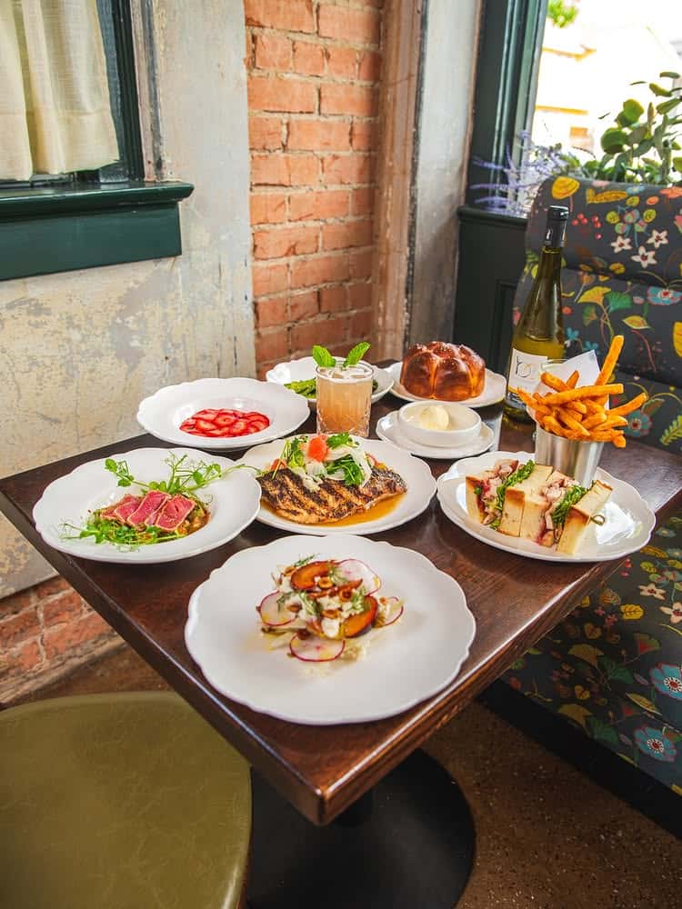 Upscale brunch table spread featuring seared ahi tuna, grilled fish with citrus salad, fresh radish and stone fruit salad, strawberry carpaccio, gourmet sandwich with sweet potato fries, house-made brioche with butter, and a bottle of white wine—set in a cozy bistro with vintage charm and floral décor.
