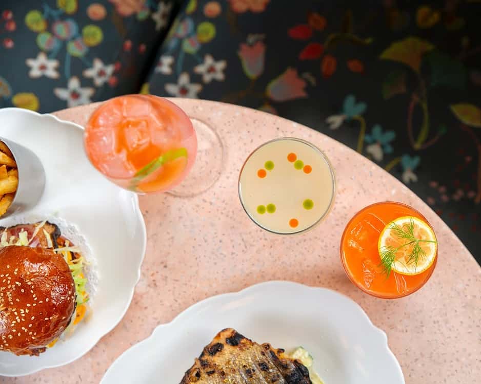 Colorful cocktails with citrus and herb garnishes served alongside a fried chicken sandwich and grilled fish at a floral-themed bistro table.