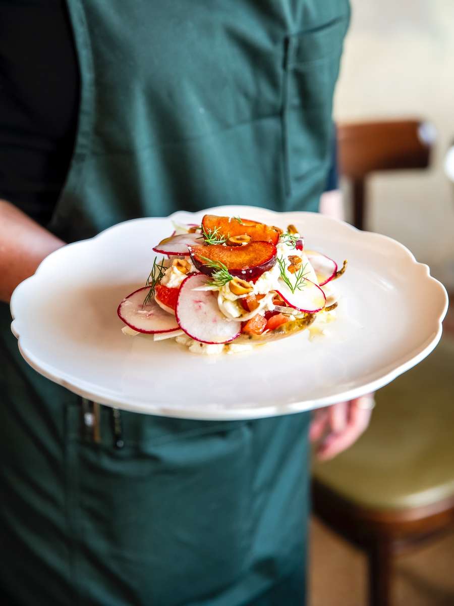 Fresh salad topped with radish slices, grilled plums, herbs, and crushed nuts, elegantly plated on a white scalloped-edge dish.